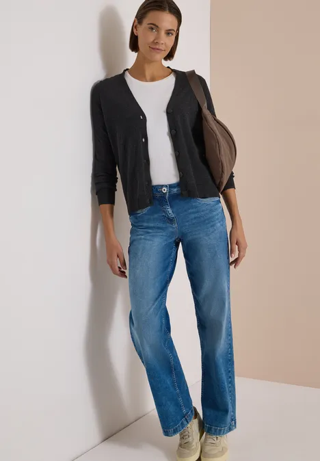 Frau lehnt an heller Wand, trägt weißes T-Shirt, schwarzen Cardigan und weite blaue Jeans.