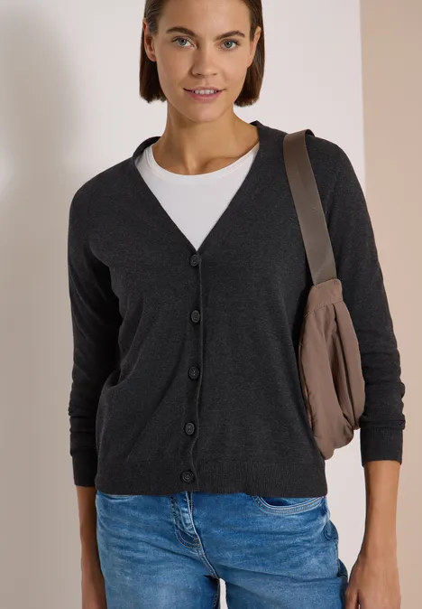 Frau trägt dunkelgrauen Cardigan, weißes Shirt, blaue Jeans und Umhängetasche vor neutralem Hintergrund.