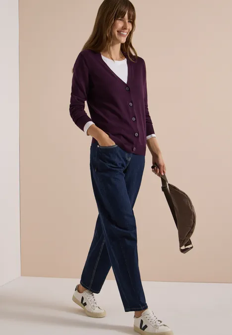 Frau trägt dunkelviolette Strickjacke, weißes Shirt, blaue Jeans, weiße Sneaker, hält Mütze.