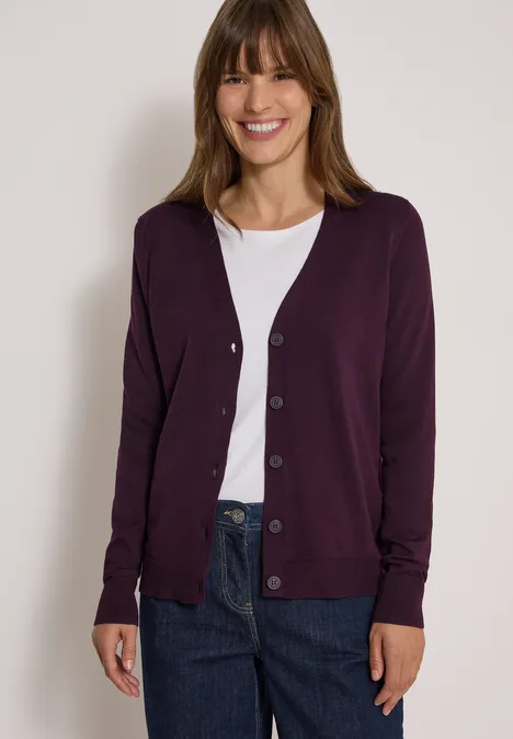 Frau mit weißem T-Shirt, lila Strickjacke und blauen Jeans lächelt vor hellgrauem Hintergrund.