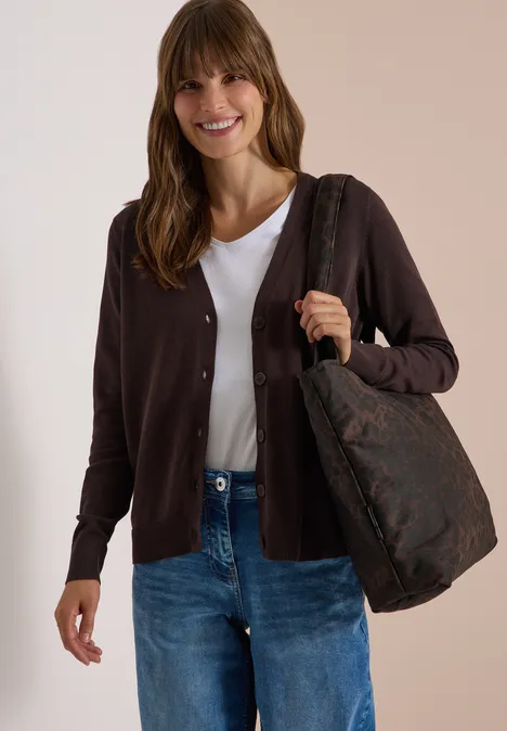 Vrouw met een donkerbruine cardigan, wit T-shirt, blauwe jeans en een grote bruine schoudertas met patroon.