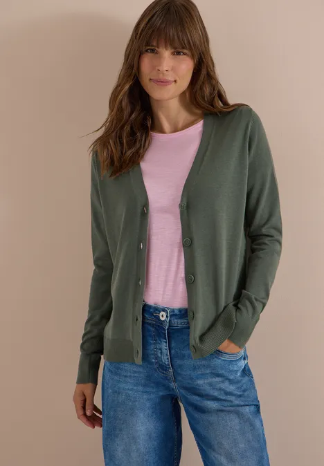 Vrouw in groene cardigan, roze shirt en jeans, handen in de zakken, voor een neutrale achtergrond.