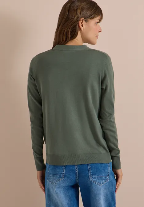Een vrouw draagt een groene trui en blauwe jeans, staat met de rug naar ons, kijkt opzij tegen een beige achtergrond.