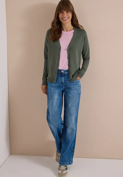 Vrouw in groen vest, roze t-shirt en wijde jeans poseert voor een beige muur.