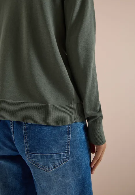 Achteraanzicht van een persoon in een groen shirt met lange mouwen en een blauwe spijkerbroek tegen neutrale achtergrond.