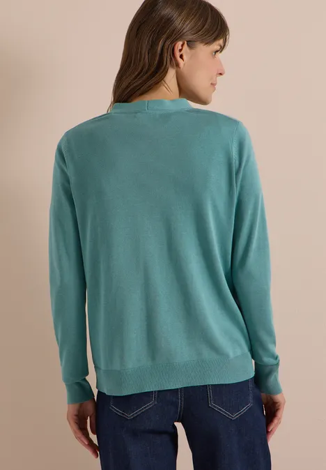 Vrouw van achteren in turquoise vest en donkere jeans voor een neutrale achtergrond.