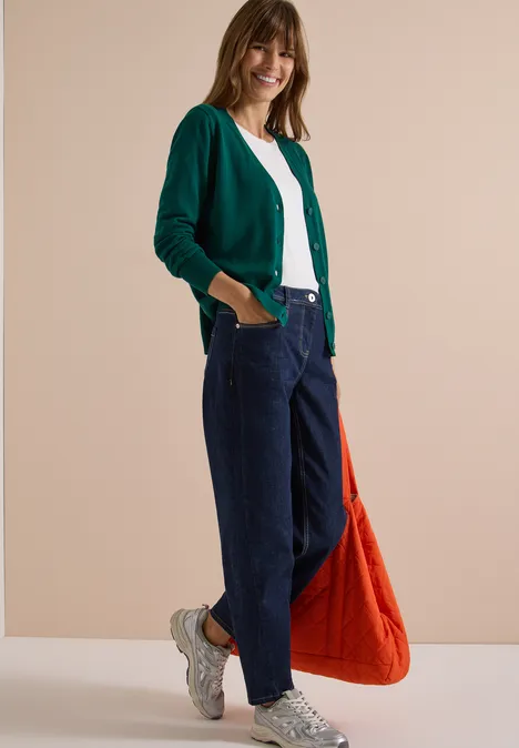 Frau in grünem Cardigan, weißem Shirt und blauen Jeans mit weißen Sneakern und orangener Tasche vor neutraler Wand.