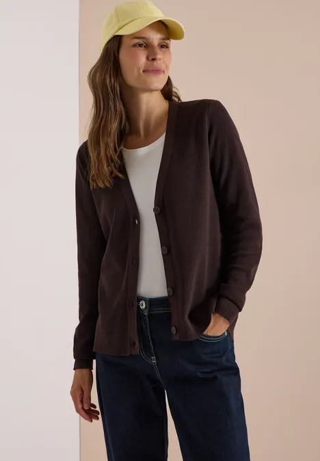 Jonge vrouw glimlachend in een bruin vest, wit T-shirt, blauwe spijkerbroek en gele pet.