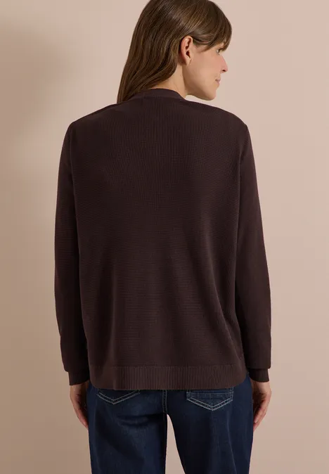 Vrouw van achteren in een donkerbruine trui en blauwe spijkerbroek tegen een beige achtergrond.