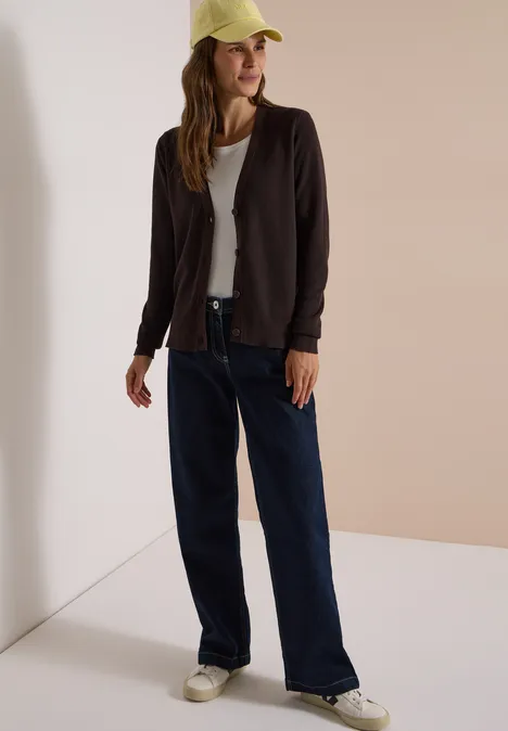Jonge vrouw in wijde jeans, wit T-shirt, donker vest en gele pet, tegen een muur leunend.