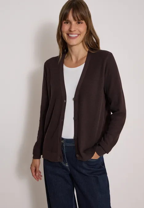 Een lachende vrouw draagt een donkerbruin vest over een wit shirt en een blauwe spijkerbroek.