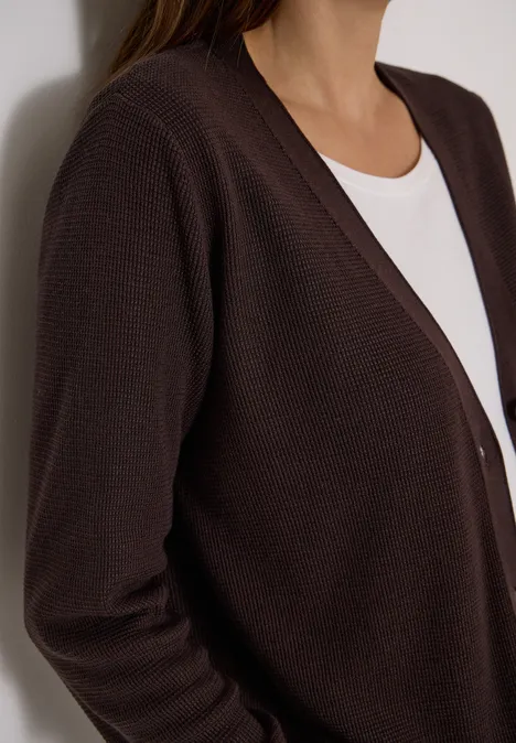 Close-up van iemand die een donkerbruin gebreid vest draagt over een wit t-shirt.