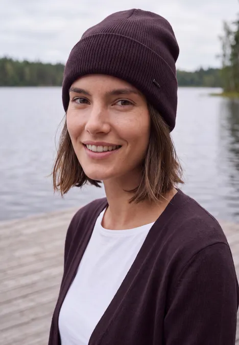 Lang openvallend vest eggplant red Junge Frau mit Mütze und Cardigan lächelt entspannt an einem Seeufer.