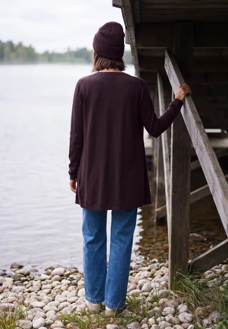 Lang openvallend vest eggplant red Rückansicht einer Person in Jeans und Strickjacke, die an einem Steg über einem See lehnt.