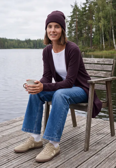 Lang openvallend vest eggplant red Frau in Jeans und Strickjacke sitzt am Holzsteg am See, hält eine Tasse und lächelt in entspannter Natur.