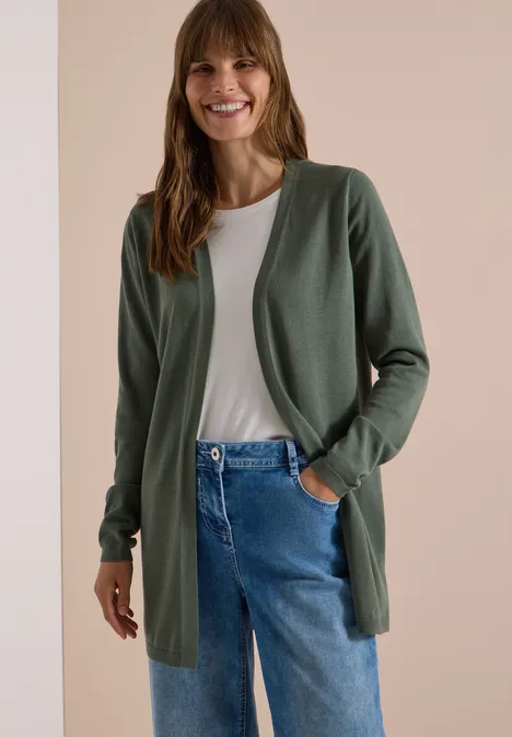 Een glimlachende vrouw in een groene cardigan, wit shirt en blauwe jeans tegen een neutrale achtergrond.