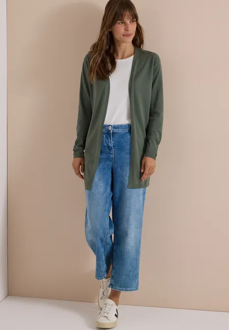 Vrouw in wit T-shirt, groen vest, wijde korte blauwe jeans en witte sneakers voor een beige muur.
