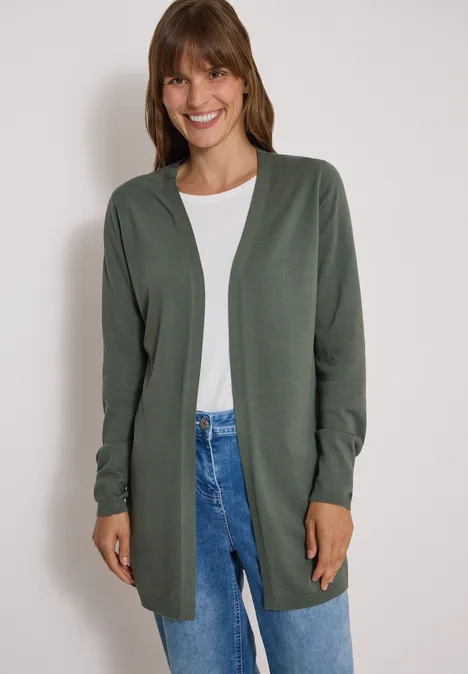 Vrouw lacht in een groene cardigan, wit shirt en spijkerbroek, voor een lichte achtergrond.