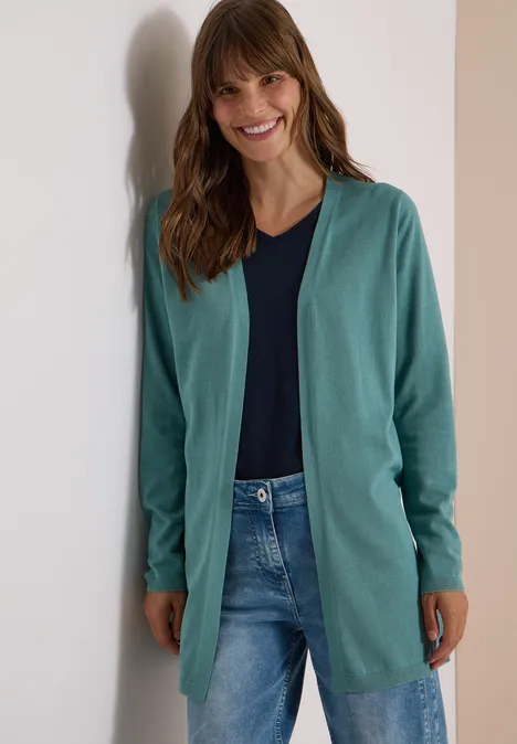 Jonge vrouw leunt glimlachend tegen een muur, draagt groene cardigan, blauw topje en spijkerbroek.