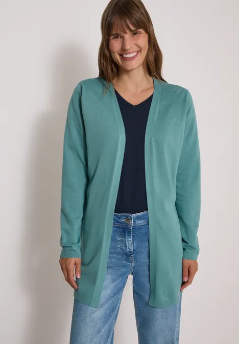 Vrouw glimlacht in groen vest, donkerblauw shirt en spijkerbroek voor lichte achtergrond.