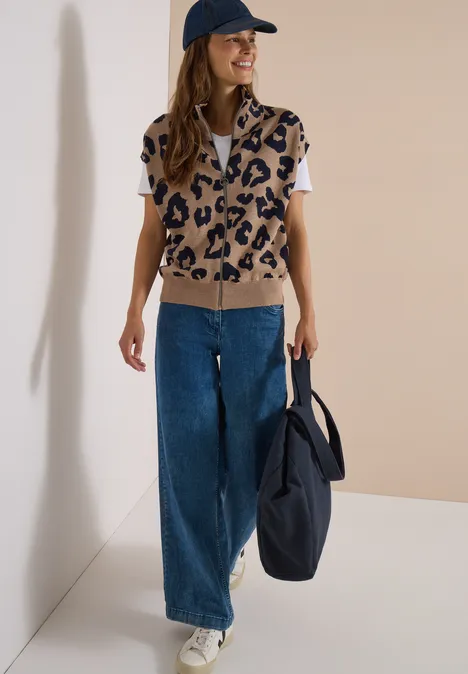 Frau trägt leo-bedrucktes Strickweste, weiße Bluse, weite Jeans, Cap, Turnschuhe und Tasche