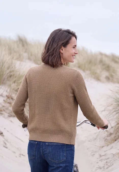 Eine Frau in braunem Pullover und Jeans schiebt am Dünenstrand ein Fahrrad, umgeben von Strandgras.