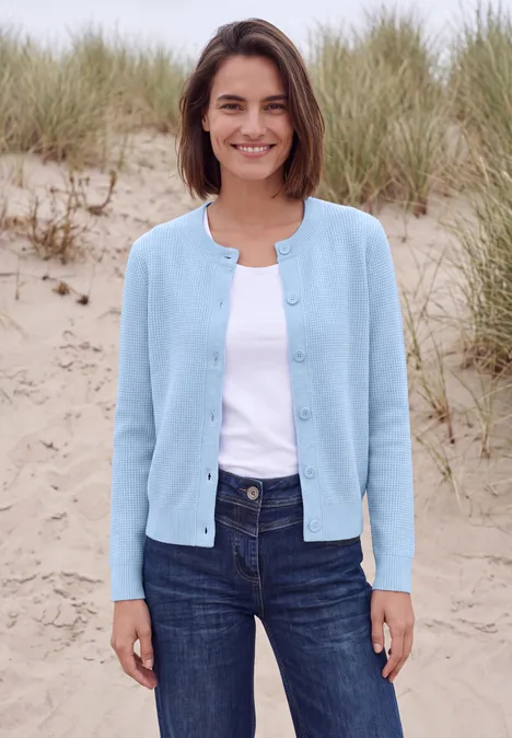 Eine lächelnde Frau in hellblauer Strickjacke, weißem Top und Jeans steht vor Sanddünen mit Gras.