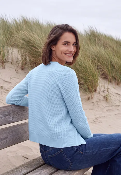 Eine Frau in einem hellblauen Pullover sitzt lächelnd auf einer Bank am Strand vor Dünen.