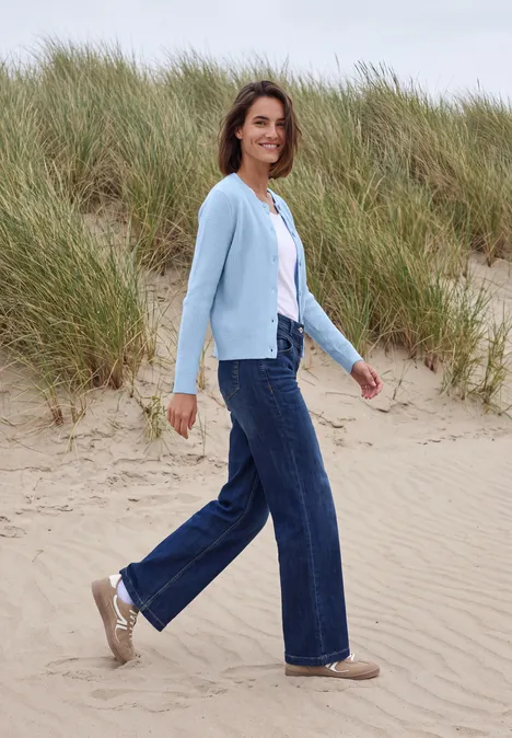 Eine Frau in Jeans, weißem Shirt und hellblauer Strickjacke spaziert über Dünen am Strand.