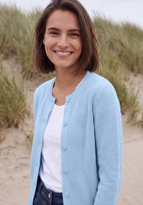 Eine lächelnde Frau in blauem Cardigan vor Dünen am Strand.