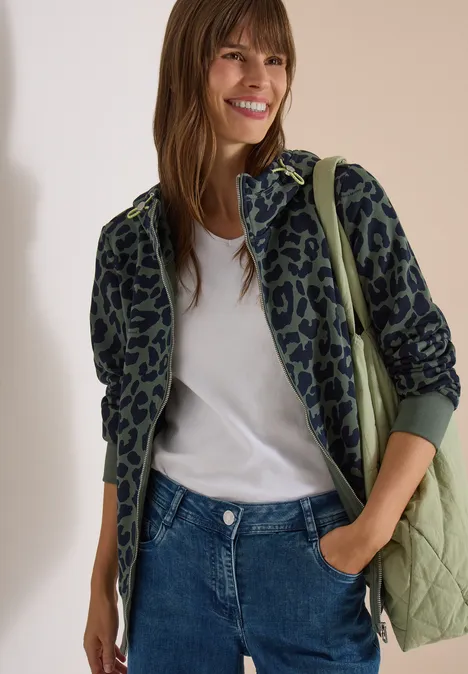 Veste de survêtement à capuche avec motif léopard dried olive green Une femme porte une veste verte à motif léopard, un t-shirt blanc, un jean et sourit.