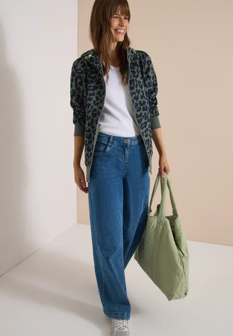 Veste de survêtement à capuche avec motif léopard dried olive green Femme souriante en veste léopard, t-shirt blanc, jean large et sac vert à la main.