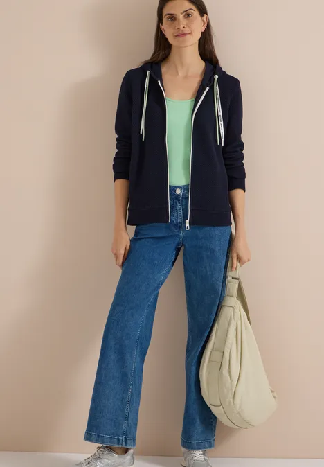 Sweatjacke mit Kapuze und Struktur urban dark blue Eine junge Frau in navyblauem Hoodie, mintgrünem Top, weiten Jeans und großer Stofftasche vor beigem Hintergrund.