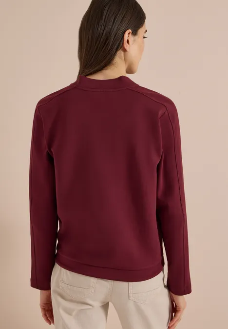 Frau von hinten in dunkelrotem Sweatshirt und hellen Hosen vor hellem Hintergrund.