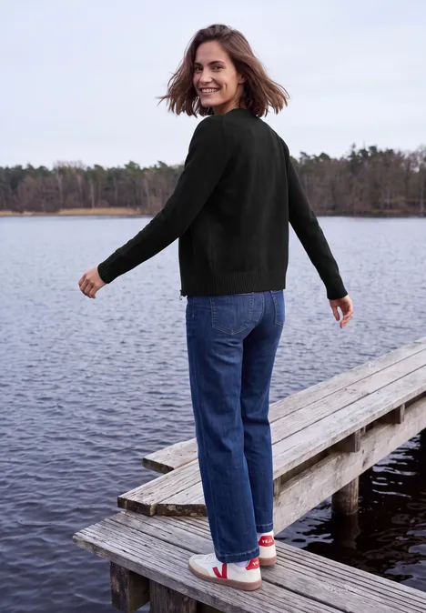 Een vrouw staat op een steiger bij een meer, kijkt glimlachend om, met zwarte trui en spijkerbroek.