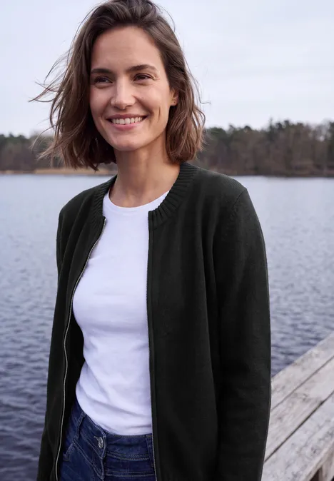 Een glimlachende vrouw in wit t-shirt en donkergroene cardigan staat op een steiger bij het meer.