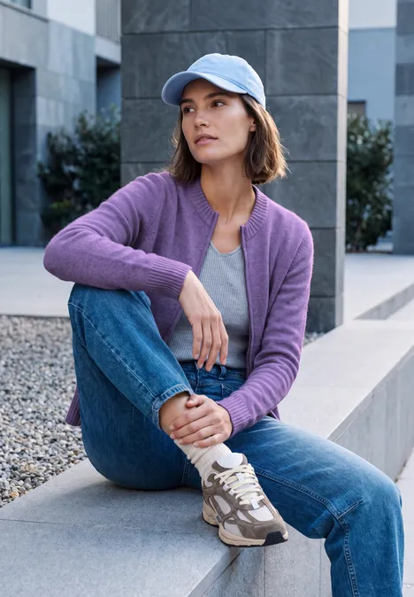 Junge Frau sitzt in lila Strickjacke, Jeans und Baseballcap auf moderner Bank im urbanen Umfeld.