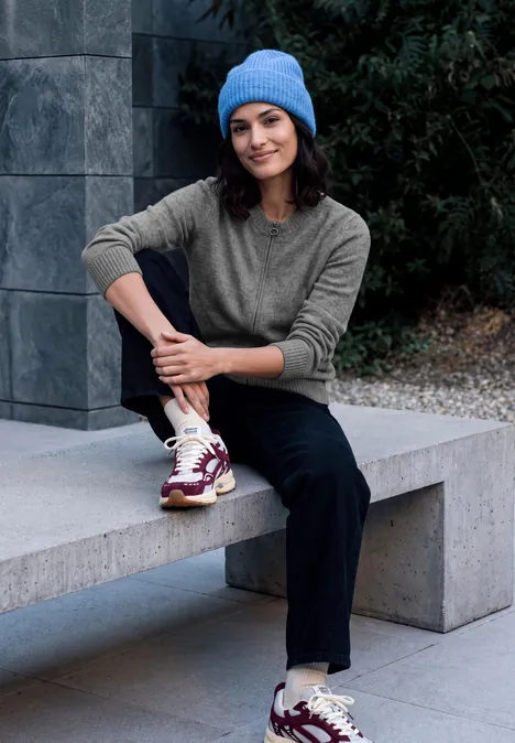 Langarm Cardigan mit Rundhals und Zipper flint grey melange Eine Person sitzt auf einer Bank mit blauer Mütze, grauem Pullover, schwarzer Hose und weißen Sneakern.