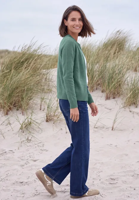 Langarm Cardigan mit Rundhals und Zipper jewel green melange Eine lächelnde Frau im grünen Pullover und Jeans steht am Strand vor Dünen mit Strandhafer.