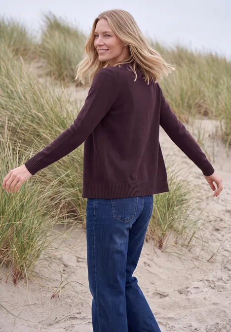 Langarm Cardigan mit Rundhals und Zipper eggplant red melange Eine blonde Frau in Pullover und Jeans spaziert am Strand entlang, Dünen im Hintergrund.