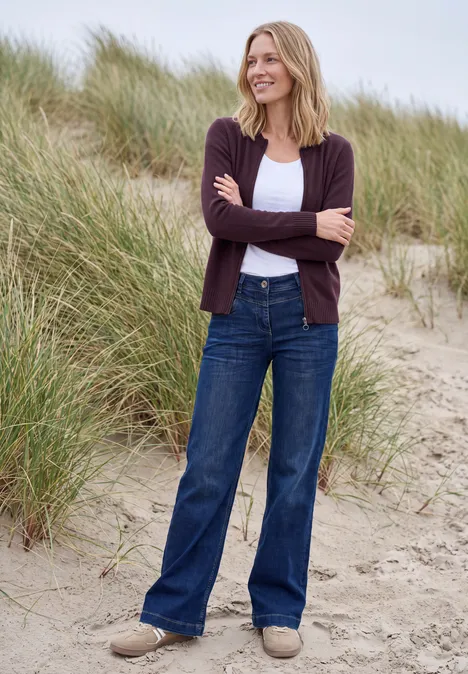 Langarm Cardigan mit Rundhals und Zipper eggplant red melange Eine lächelnde Frau steht in Dünen am Strand, trägt braune Strickjacke, weißes Top, weite Jeans und helle Schuhe.