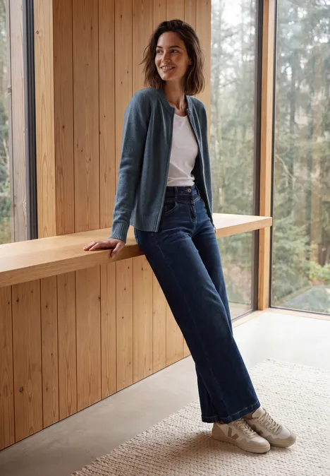 Vrouw in jeans en vest leunt ontspannen op een houten bank in een moderne, lichtdoorlatende ruimte.