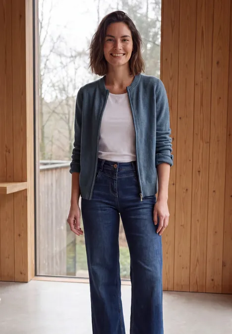 Jonge vrouw in jeans en wit shirt, lachend en ontspannen, staande voor houten panelen en een raam.