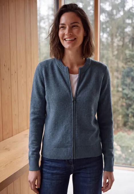 Een glimlachende vrouw in een blauw vest met rits en spijkerbroek staat binnen voor een houten wand en een raam.