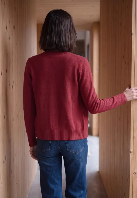 Eine Person steht mit dem Rücken zur Kamera in einem Holzflur, trägt roten Pullover und blaue Jeans.