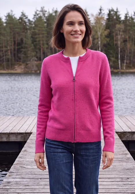 Langarm Cardigan mit Rundhals und Zipper pink flash Frau steht auf einem Holzsteg am See, trägt pinke Strickjacke und Jeans, im Hintergrund Waldseenlandschaft.
