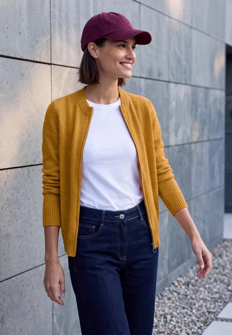 Cardigan zippé à manches longues et col rond golden honey melange Frau in gelbem Cardigan, weißem T-Shirt und Jeans mit roter Kappe spaziert lächelnd an einer Steinmauer entlang.