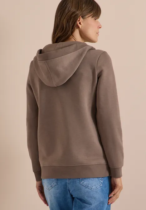 Rückansicht einer Frau in brauner Kapuzenjacke und blauen Jeans vor hellem Hintergrund.