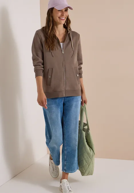 Junge Frau in brauner Kapuzenjacke, blauen Jeans, weißen Sneakern und grüner Tasche vor neutralem Hintergrund.