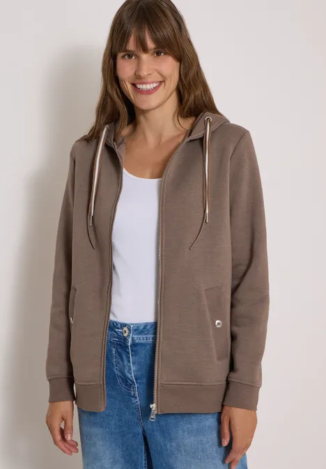 Frau in brauner Kapuzenjacke, weißem Top und Jeans, lächelt vor hellem Hintergrund.